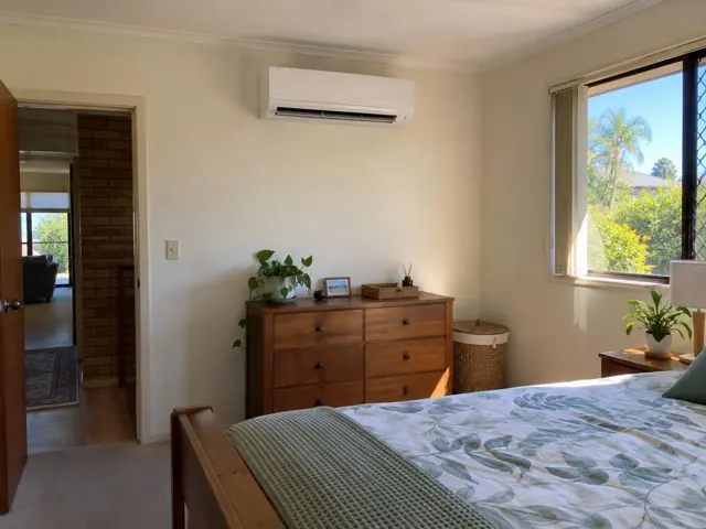 Wall-mounted split system above a dresser in a bright bedroom with window and timber furniture in Coombabah